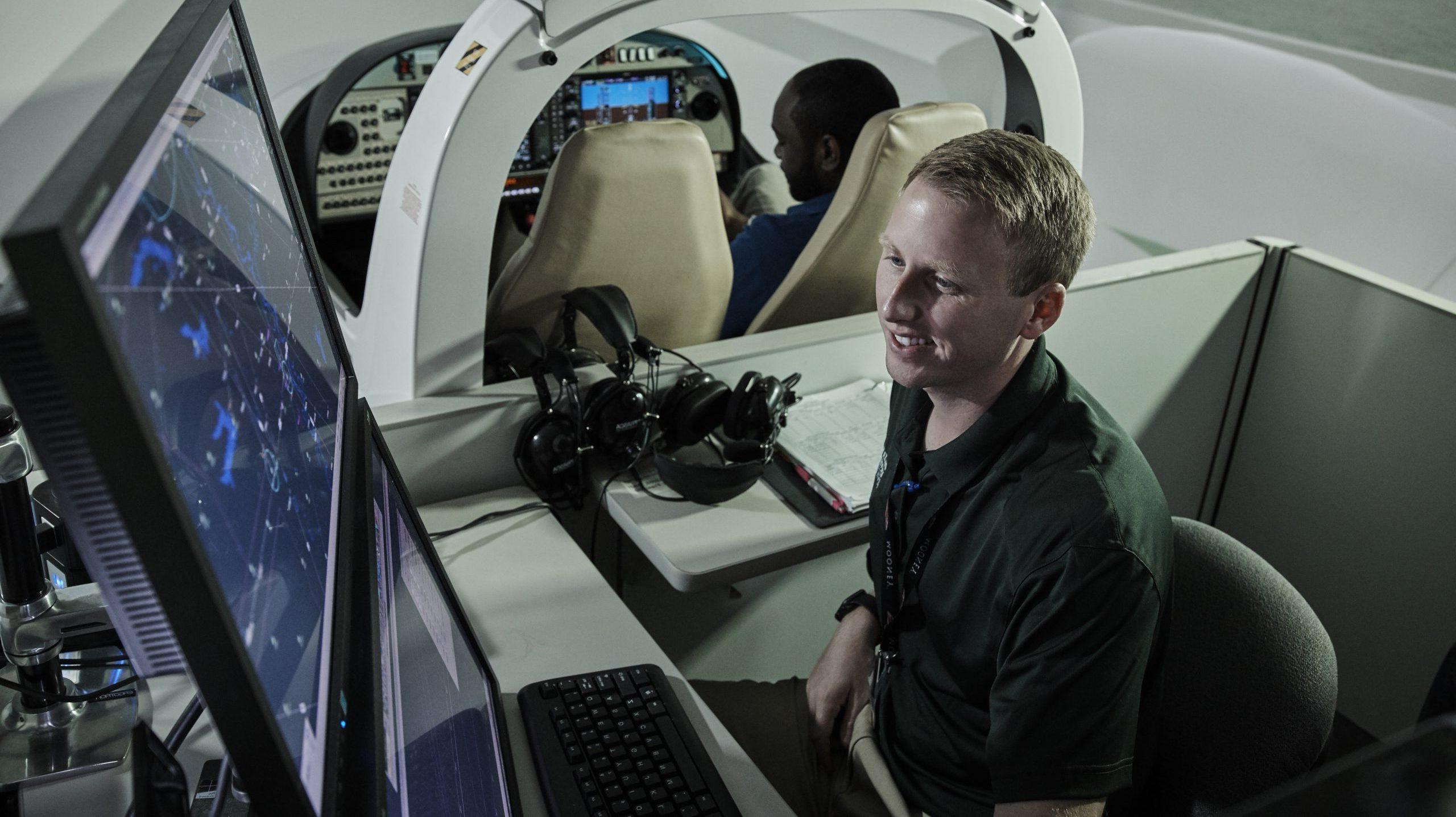 看着电脑屏幕的学生, 监察空中交通, while another student is in the flight simulator behind him.